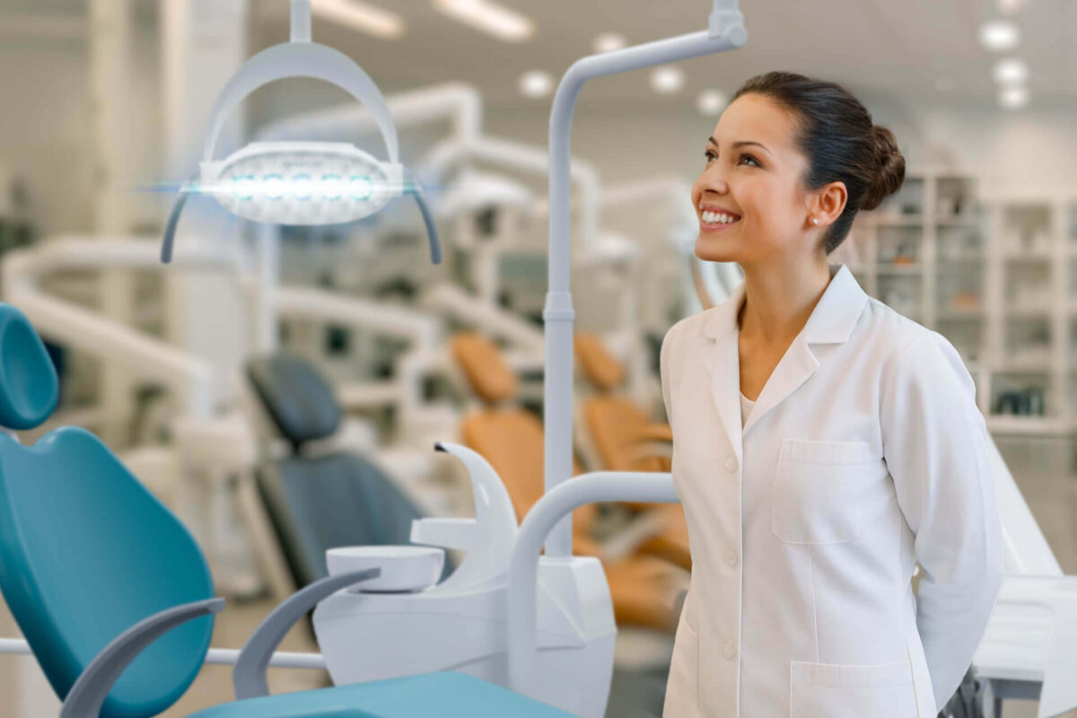 Qual a melhor cadeira odontológica? Saiba como acertar na escolha! gerente de consultório odontológico Dentista sorridente em consultório moderno observando o equipamento de iluminação ao lado da melhor cadeira odontológica, destacando conforto e tecnologia