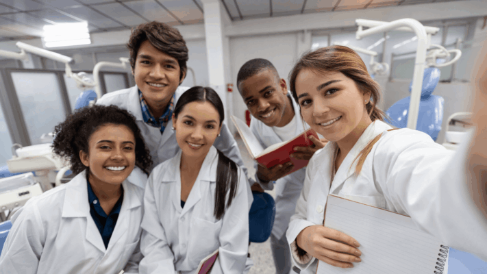 Estudantes em uma faculdade de odontologia sorrindo durante aula prática em um laboratório com cadeiras odontológicas ao fundo