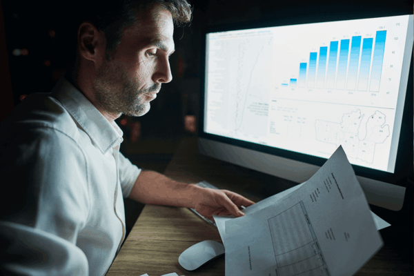 Homem analisando relatórios e gráficos de desempenho no computador, avaliando dados financeiros relacionados a franquias em um ambiente de trabalho noturno.